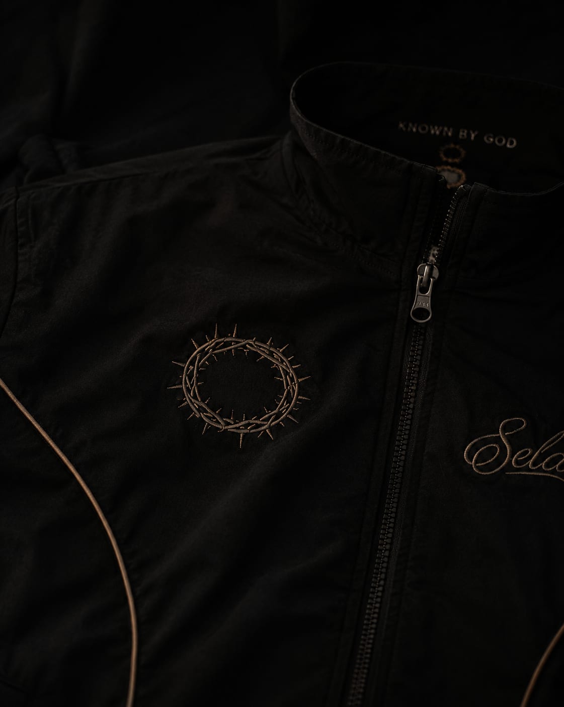 Stillness Track Jacket in black — tonal crown of thorns embroidery, KNOWN BY GOD collar, leather-brown piping